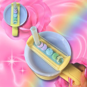 May include: A yellow and white reusable cup with a lid that has a built-in straw holder and a removable straw. The lid has a glitter design and pastel-colored moon shapes.