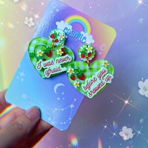 Peut inclure: Une paire de boucles d'oreilles en forme de cœur en vichy vert et blanc avec un motif de fraises et de fleurs. La boucle d'oreille gauche dit "J'ai jamais eu peur" et la boucle d'oreille droite dit "avant que tu n'arrives".