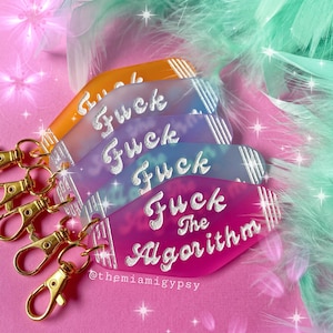 May include: A set of five colorful keychains with the words "Fuck" and "Fuck the Algorithm" written in white on them. The keychains are arranged in a stack, with the top keychain being pink and the bottom keychain being orange. The keychains are on a pink background with white glitter.