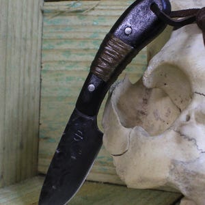 May include: A small, handcrafted black knife with a wrapped handle and a leather lanyard. The blade has a curved edge and a dark finish. The handle is secured with silver rivets. The knife is displayed against a wooden background.