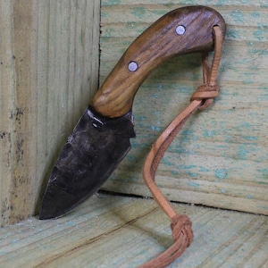 Peut inclure: Un petit couteau artisanal avec une lame sombre et texturée et un manche en bois. La poignée est fixée avec deux rivets argentés et comporte une lanière en cuir. Le couteau est conçu pour l'utilité et l'utilisation en extérieur.