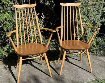 Könnte beinhalten: Zwei Holzstühle mit hellbrauner Oberfläche und einer Lattenrücklehne und einem Latten-Sitz. Die Stühle haben ein schlichtes Design mit abgewinkelten Beinen und Armlehnen.