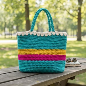 Può includere: Una borsa tote all'uncinetto turchese con un design a righe gialle e rosa. La borsa ha un bordo smerlato e manici turchesi. La borsa è appoggiata su un tavolo da picnic in legno.