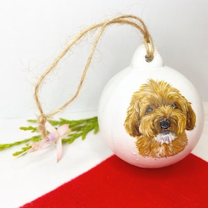 May include: A white ceramic Christmas ornament with a hand-painted portrait of a brown and white dog. The ornament has a twine hanger.