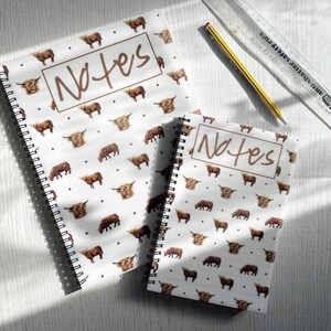 May include: Two spiral-bound notebooks with a white background and a pattern of brown Highland cows. The word "Notes" is written in brown script on the front cover. A pencil and ruler are also visible.