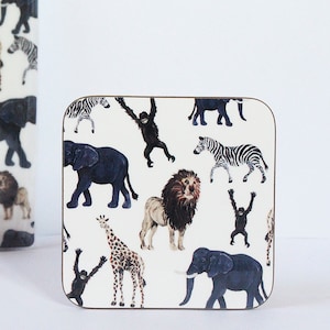 May include: A square coaster with a white background and a repeating pattern of black and white zebras, black elephants, brown giraffes, and brown lions. The coaster has a brown wooden frame.