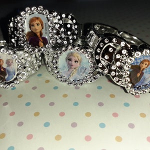 May include: Four silver plastic rings with a jeweled design. Each ring features a different image of a Disney Frozen character. The rings are on a white background with pink, blue, and yellow polka dots.