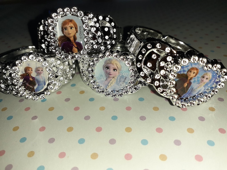 May include: Four silver plastic rings with a jeweled design. Each ring features a different image of a Disney Frozen character. The rings are on a white background with pink, blue, and yellow polka dots.