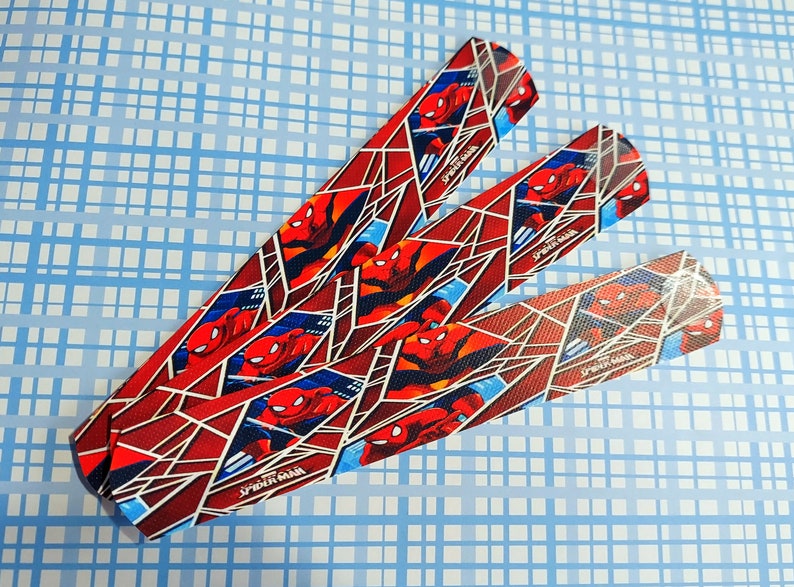 Peut inclure: Trois bandes de tissu rouge et bleu avec un motif Spider-Man. Les bandes ont un fond blanc avec un motif de toile rouge et des images de Spider-Man. Le texte "Spider-Man" est imprim&eacute; sur chaque bande.