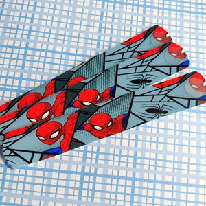 Peut inclure: Trois bandes de tissu bleu et gris &agrave; motifs avec une image de style bande dessin&eacute;e de Spiderman en rouge et bleu. Les bandes sont superpos&eacute;es.