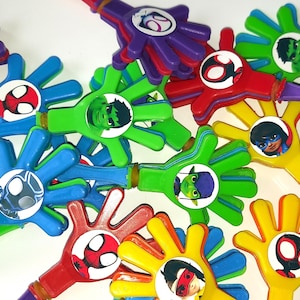 Peut inclure: Un assortiment de claquettes en forme de main aux couleurs vives, chacune représentant un super-héros différent. Les claquettes sont bleues, vertes, rouges, jaunes et violettes, avec des images de personnages au centre. Chaque claquette a une poignée et un élastique.