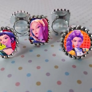 May include: Silver-toned adjustable rings with colorful images of animated characters. The rings have a scalloped edge and are set on a polka-dot surface. The images feature women with various hairstyles and outfits.