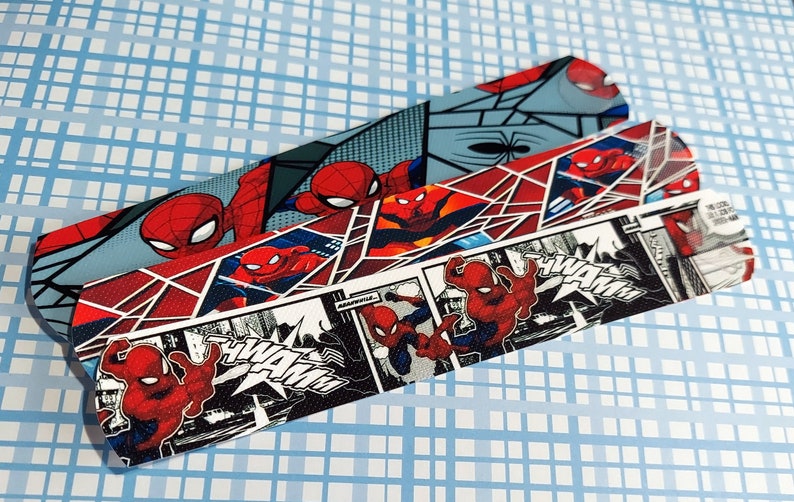 Peut inclure: Trois bandes de tissu rouge, noir et blanc avec un motif de bande dessin&eacute;e mettant en sc&egrave;ne Spiderman. Les bandes pr&eacute;sentent un motif r&eacute;p&eacute;t&eacute; de Spiderman dans diff&eacute;rentes poses et les mots "Wham!" et "Pow!" dans une police de caract&egrave;res de bande dessin&eacute;e.