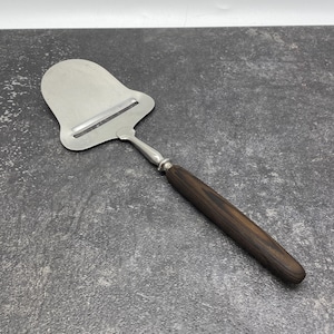May include: A cheese slicer with a stainless steel blade and a dark brown wooden handle. The blade has a rectangular slot for slicing. The handle is cylindrical and made of dark wood. The slicer is designed for cutting cheese.