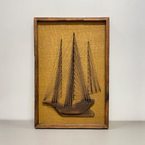 Peut inclure: Une œuvre d'art en fil encadrée représentant un voilier à trois mâts. Le bateau est fait de bois foncé et de fil noir, sur un fond de toile de jute beige. Le cadre en bois ajoute une touche rustique à cette œuvre d'art sur le thème nautique.