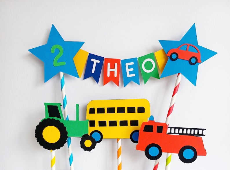 May include: A colorful birthday cake topper featuring a tractor, school bus, and fire truck. The topper includes blue star accents with the number 2 and a banner that spells out "THEO". The vehicles are green, yellow, and red.