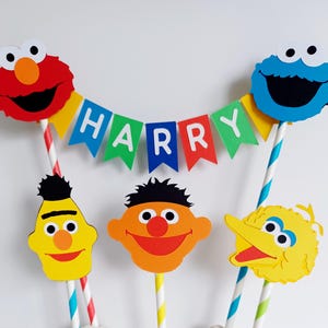 May include: A Sesame Street themed cake topper set with Elmo, Cookie Monster, Big Bird, and Bert. The set includes a banner that says "HARRY".