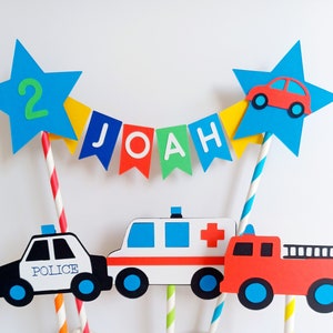 Primary coloured bunting cake topper with stripy straws.  2 stars either side of the bunting displaying the age in one star and a car in the other.  This topper comes complete with 3 vehicles.  This image shows a police car, fire engine & ambulance.
