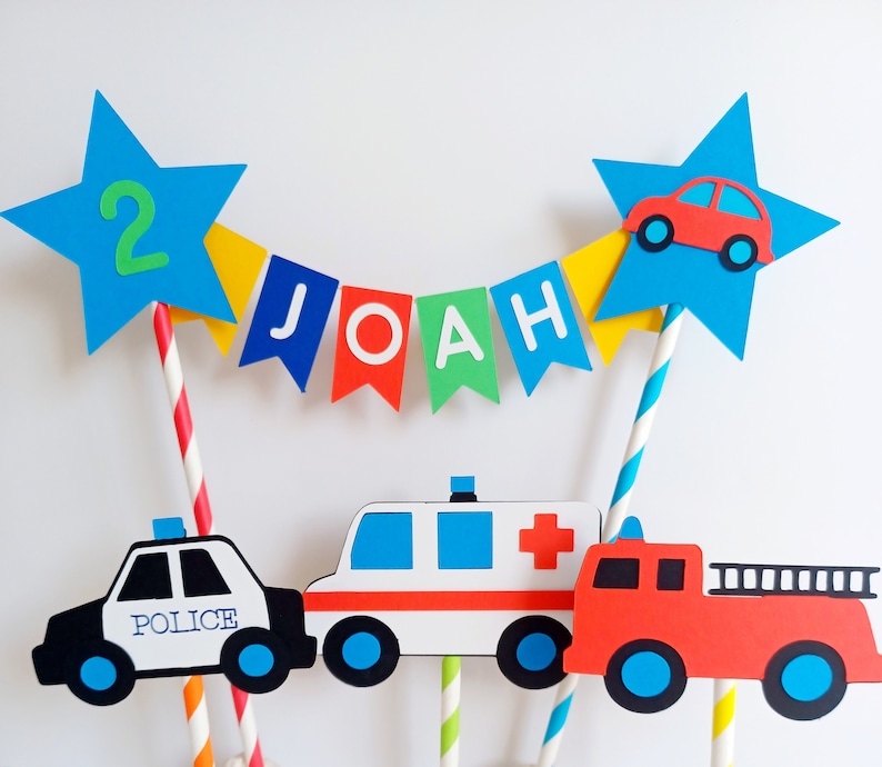 Primary coloured bunting cake topper with stripy straws.  2 stars either side of the bunting displaying the age in one star and a car in the other.  This topper comes complete with 3 vehicles.  This image shows a police car, fire engine & ambulance.