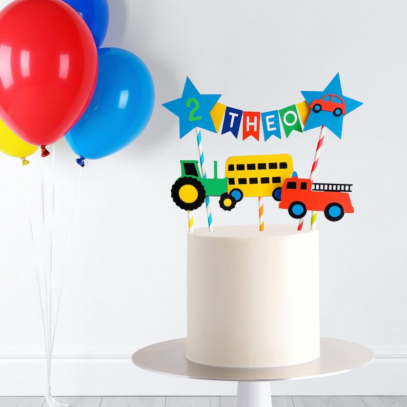 May include: A white cake with colorful toppers, including a green tractor, yellow school bus, and red fire truck. A banner spells "THEO" and a blue star displays the number "2". Balloons in red, blue, and yellow are in the background.