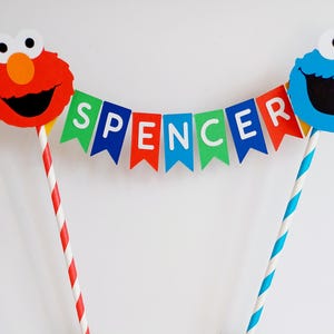 May include: A cake topper featuring Elmo and Cookie Monster with a banner that reads "Spencer". The banner is made of colorful paper flags with white letters. The topper is perfect for a birthday party.