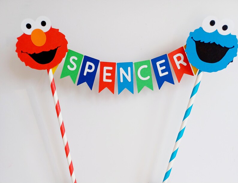 May include: A cake topper featuring Elmo and Cookie Monster with a banner that reads "Spencer". The banner is made of colorful paper flags with white letters. The topper is perfect for a birthday party.