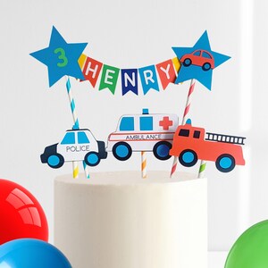 May include: Cake topper featuring a police car, ambulance, and fire truck. The topper includes blue star accents with the number 3 and a banner that spells out "HENRY". The cake topper is designed for a birthday celebration.