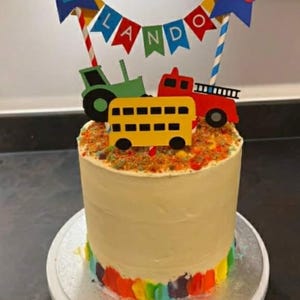 May include: A birthday cake decorated with a colorful rainbow base and topped with a variety of vehicle cutouts. The cake is adorned with a banner that spells out a name and a star with the number 2.
