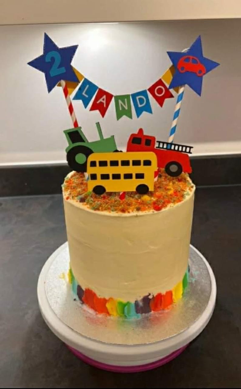 May include: A birthday cake decorated with a colorful rainbow base and topped with a variety of vehicle cutouts. The cake is adorned with a banner that spells out a name and a star with the number 2.