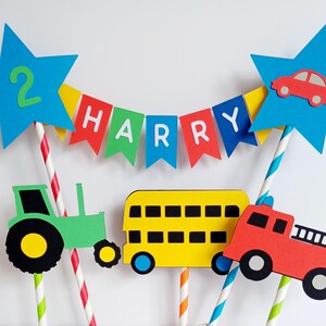 May include: A colorful birthday cake topper featuring a blue star with the number 2, a banner that spells out "HARRY", and various vehicle cutouts including a tractor, bus, and fire truck. The topper is designed with paper straws.