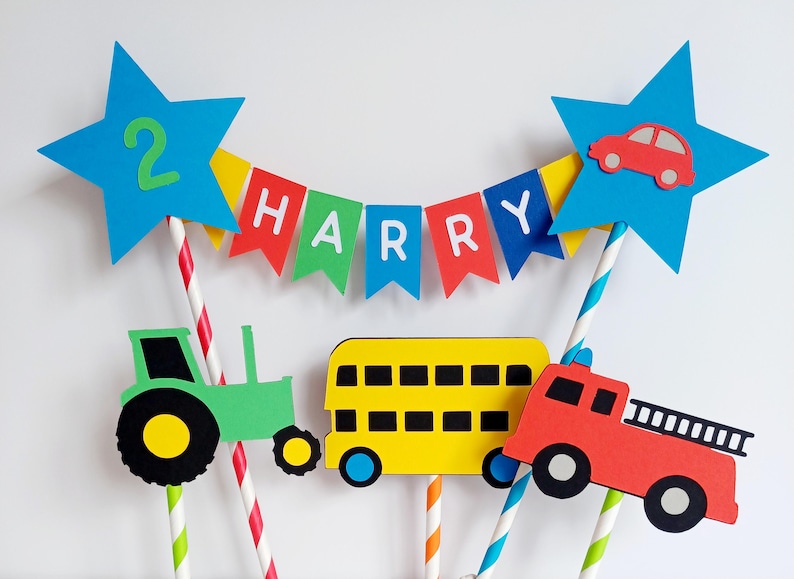 May include: A colorful birthday cake topper featuring a blue star with the number 2, a banner that spells out "HARRY", and various vehicle cutouts including a tractor, bus, and fire truck. The topper is designed with paper straws.