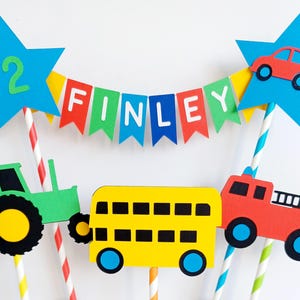 May include: A colorful birthday cake topper featuring a banner that spells "FINLEY" and a blue star with the number "2". The topper includes cutouts of a red car, a green tractor, a yellow school bus, and a red fire truck.