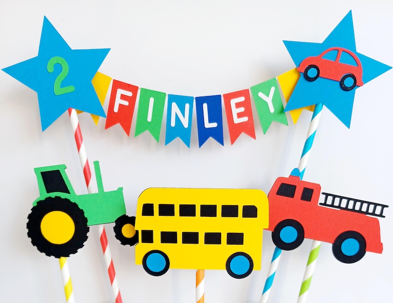May include: A colorful birthday cake topper featuring a banner that spells "FINLEY" and a blue star with the number "2". The topper includes cutouts of a red car, a green tractor, a yellow school bus, and a red fire truck.