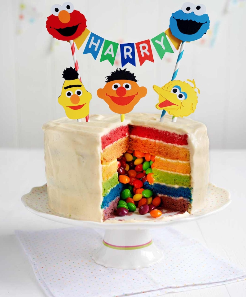 May include: A rainbow layered cake with a white frosting and a Sesame Street theme. The cake is decorated with a banner that says "HARRY" and Sesame Street character toppers. The cake is filled with colorful candies.
