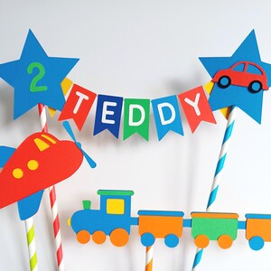 May include: A colorful cake topper featuring a red airplane, a blue train with orange and green cars, and a banner that spells "TEDDY". Two blue star-shaped toppers hold a red car and the number "2". The cake topper is designed for a birthday.