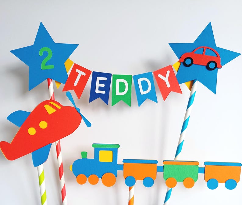 May include: A colorful cake topper featuring a red airplane, a blue train with orange and green cars, and a banner that spells "TEDDY". Two blue star-shaped toppers hold a red car and the number "2". The cake topper is designed for a birthday.