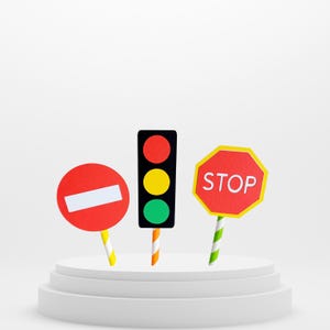 May include: Three traffic signs on a white platform. A red circle with a white bar, a black traffic light with red, yellow, and green lights, and a red octagon with the word "STOP" in white. Each sign has a striped pole.