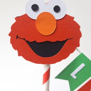 May include: Red and orange felt Elmo cake topper with a black smile and white eyes. The topper is attached to a red and white striped paper straw.