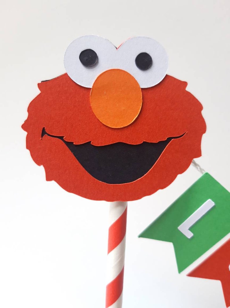 May include: Red and orange felt Elmo cake topper with a black smile and white eyes. The topper is attached to a red and white striped paper straw.