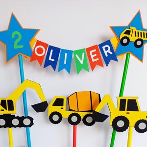 May include: A colorful construction-themed birthday cake topper set. Includes yellow construction vehicles, blue star toppers with the number 2 and a dump truck, and a banner spelling "OLIVER".