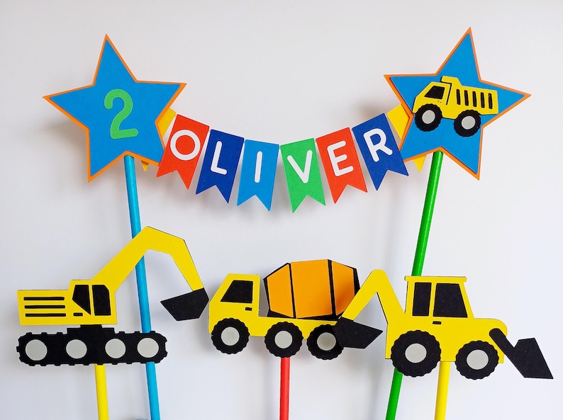 May include: A colorful construction-themed birthday cake topper set. Includes yellow construction vehicles, blue star toppers with the number 2 and a dump truck, and a banner spelling "OLIVER".