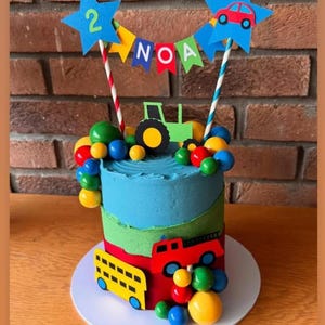 May include: A birthday cake decorated with blue, green, and red frosting. The cake features a green tractor, a red car, and a yellow bus. The cake is topped with blue star-shaped decorations with the number 2 and the name "NOA". Colorful candies surround the cake.