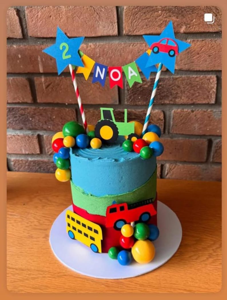 May include: A birthday cake decorated with blue, green, and red frosting. The cake features a green tractor, a red car, and a yellow bus. The cake is topped with blue star-shaped decorations with the number 2 and the name "NOA". Colorful candies surround the cake.
