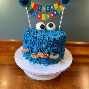 May include: A blue and white Cookie Monster themed cake with a blue frosting and chocolate chip cookie decorations. The cake is decorated with a banner that says "Zack is one".