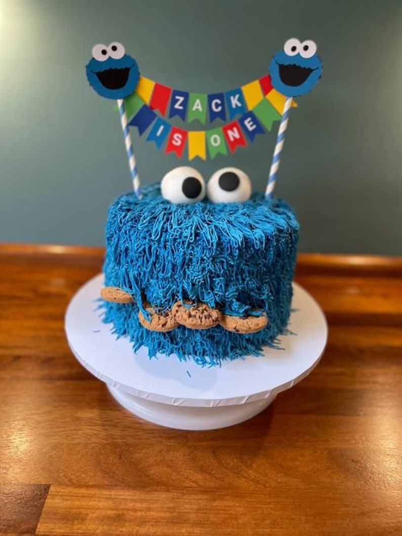 May include: A blue and white Cookie Monster themed cake with a blue frosting and chocolate chip cookie decorations. The cake is decorated with a banner that says "Zack is one".