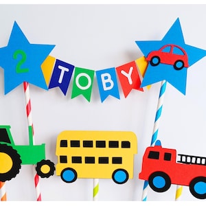 May include: A colorful cake topper featuring a blue star with the number 2, a banner spelling "TOBY", and vehicle cutouts including a green tractor, yellow school bus, red fire truck, and a red car. Perfect for a birthday celebration.
