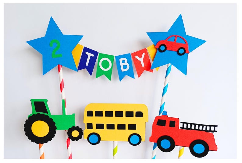 May include: A colorful cake topper featuring a blue star with the number 2, a banner spelling "TOBY", and vehicle cutouts including a green tractor, yellow school bus, red fire truck, and a red car. Perfect for a birthday celebration.