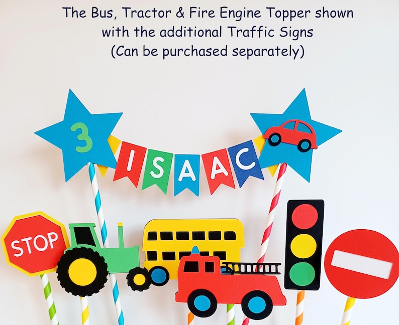 May include: Colorful birthday cake toppers featuring a bus, tractor, and fire engine. The toppers include a banner spelling "ISAAC", a star with the number "3", and various traffic signs. The text "The Bus, Tractor & Fire Engine Topper shown with the additional Traffic Signs (Can be purchased separately)" is also visible.