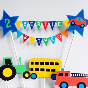 May include: A colorful cake topper with a blue star featuring the number 2, a banner reading "BERTIE & THEO", and cutouts of a car, tractor, bus, and fire truck. The topper is designed for a birthday celebration.