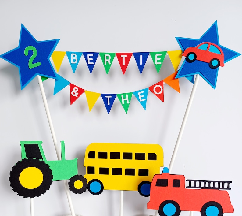 May include: A colorful cake topper with a blue star featuring the number 2, a banner reading "BERTIE & THEO", and cutouts of a car, tractor, bus, and fire truck. The topper is designed for a birthday celebration.